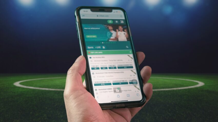 A Person Holding a Smartphone Displaying a Sports Betting Website, with A Blurred Background of A Soccer Field Under Stadium Lights