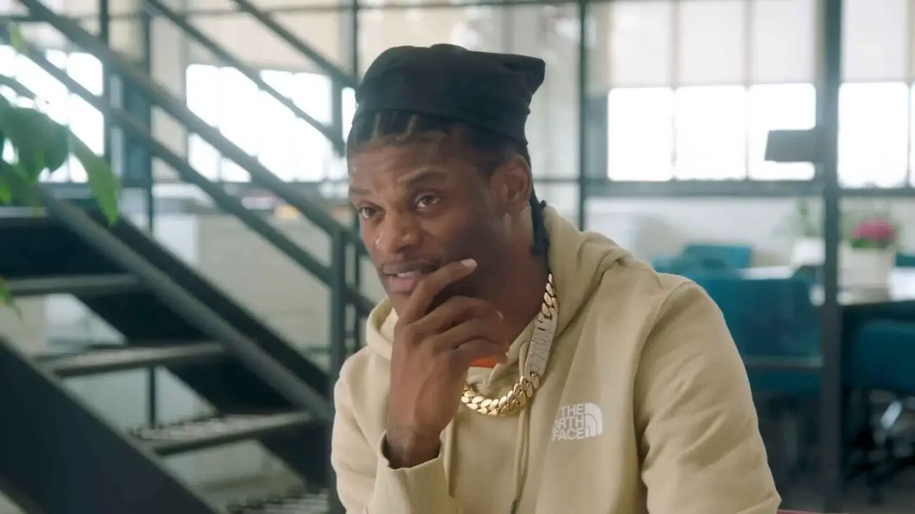 Lamar Jackson wearing a beige hoodie and gold chain, seated indoors during an interview, resting his hand on his chin