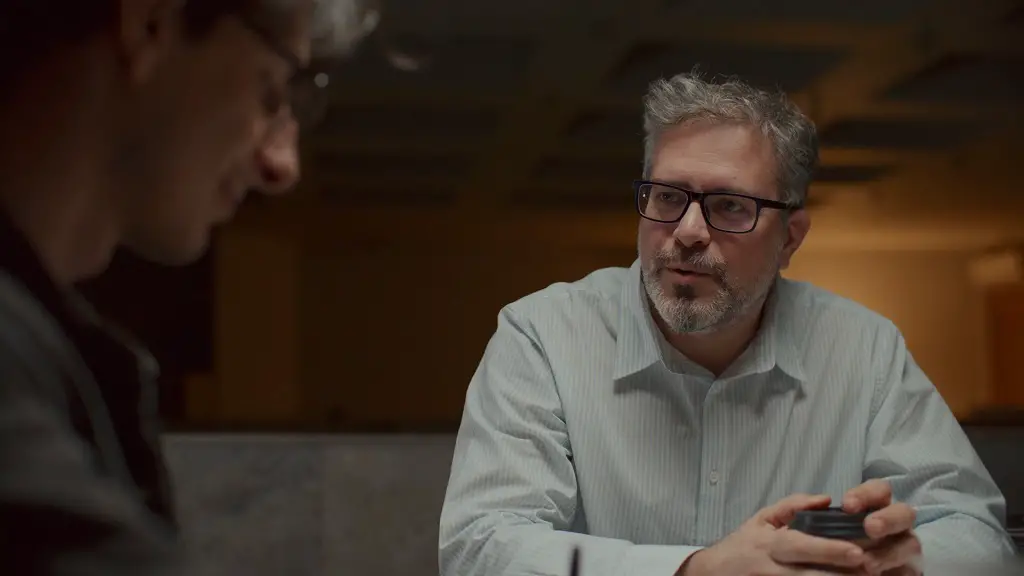 A middle-aged man wearing glasses speaks seriously to another person across a table