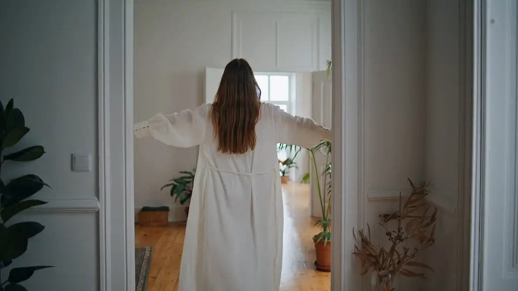 A woman in a white robe walks through a bright hallway with her arms slightly raised