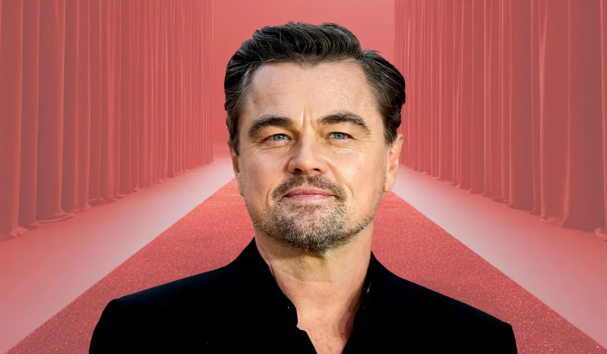 A person in a black suit standing on a red carpet with curtains in the background