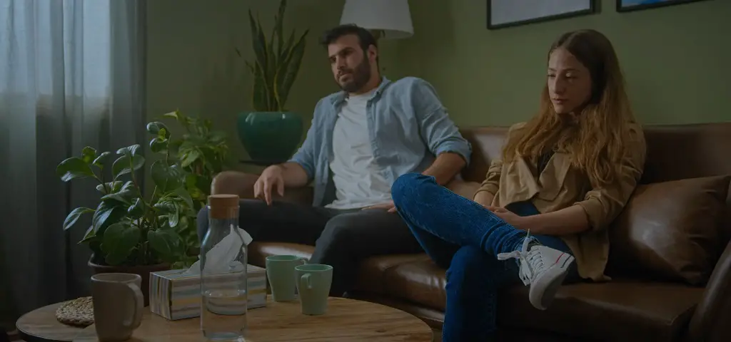 A man and woman sit tensely on a living-room sofa, avoiding eye contact during a difficult conversation
