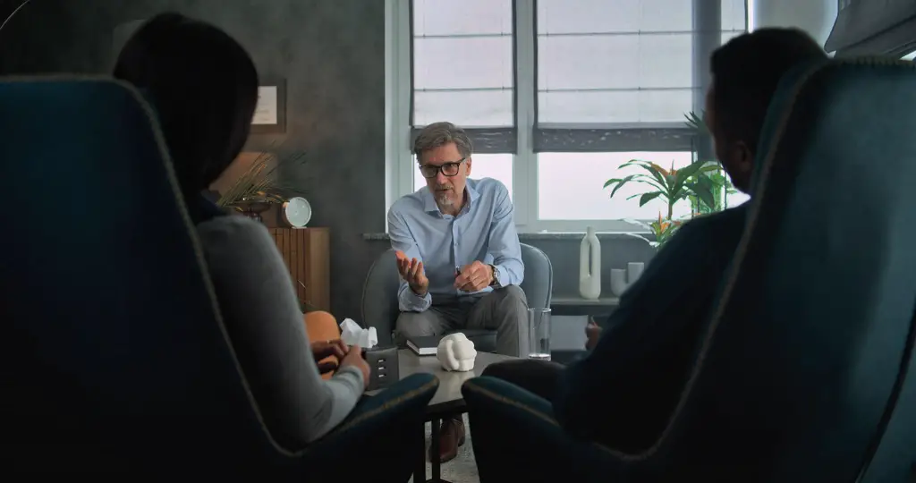 A counselor speaks to two individuals seated across from him in a therapy office