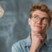 Thoughtful young man wearing glasses looking upward beside a glowing light bulb