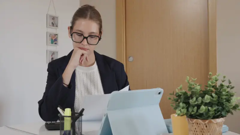 A woman reviews paperwork at her desk with a tablet open beside her