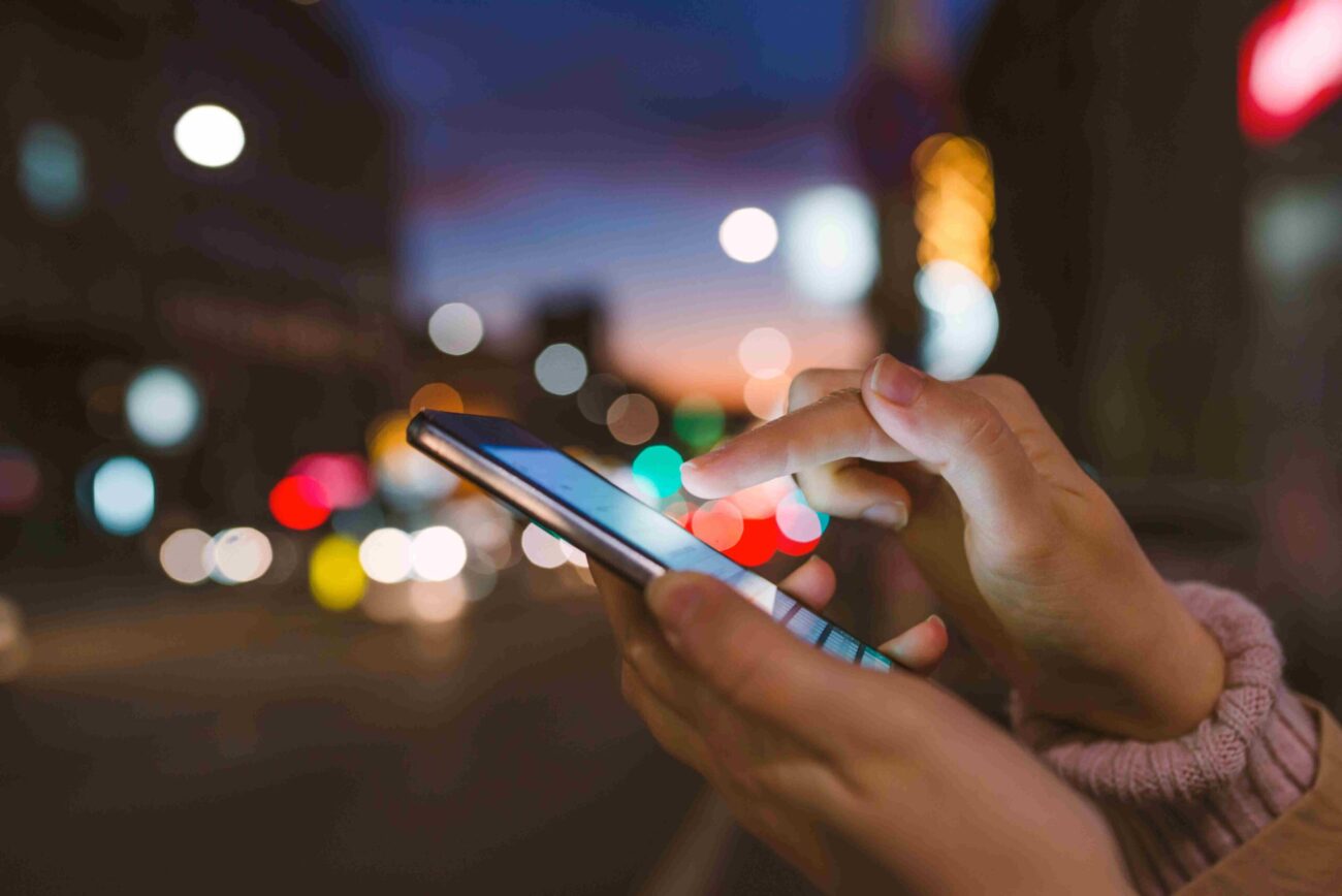 Close-up of hands using a smartphone at night with colorful city lights blurred in the background