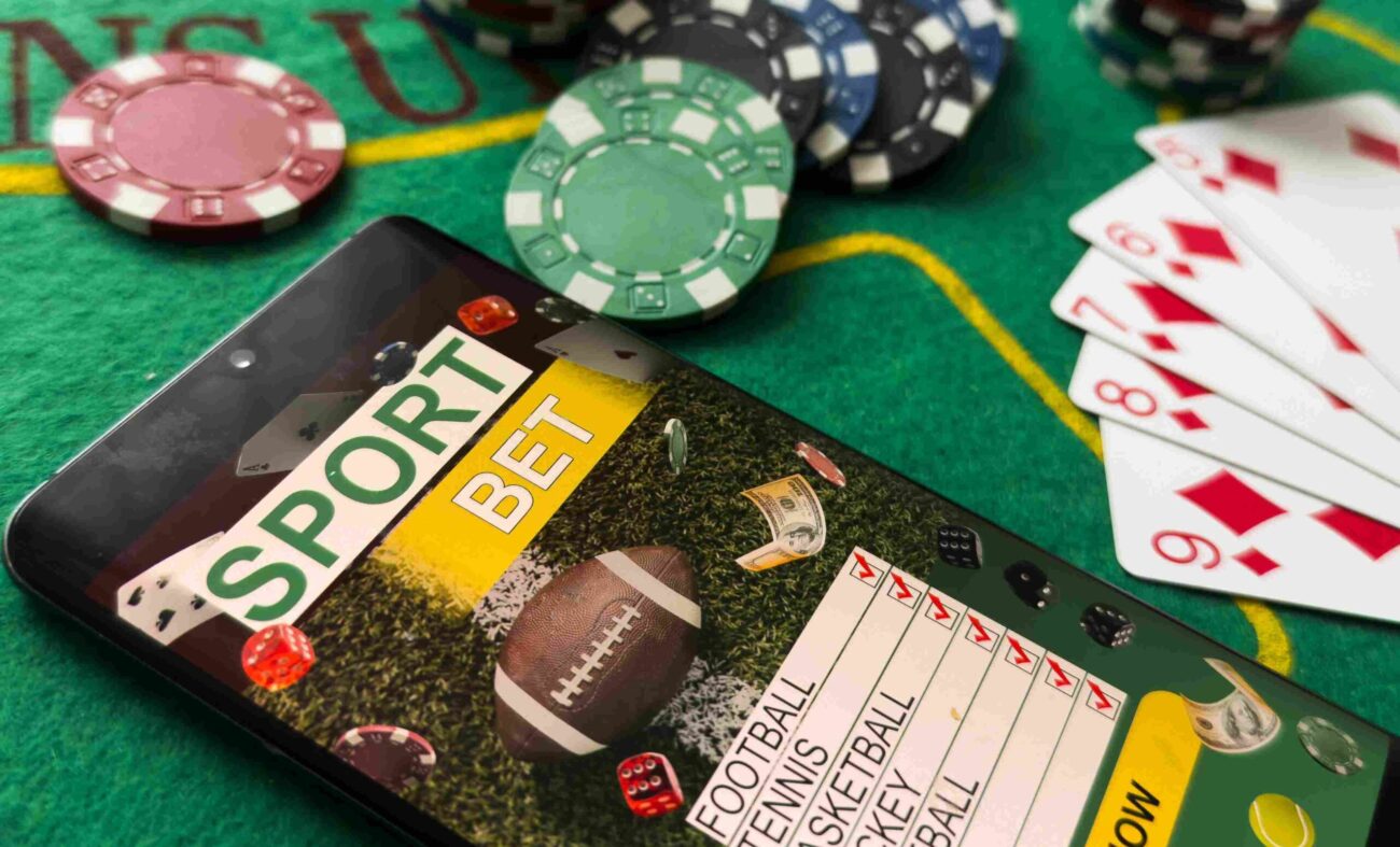 Smartphone displaying a sports betting app on a casino table beside poker chips and playing cards