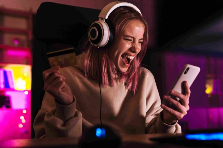 Excited woman wearing headphones holding a credit card and looking at her smartphone in a colorful gaming setup