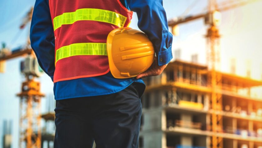Construction worker holds a hard hat near an active building site with cranes in the background