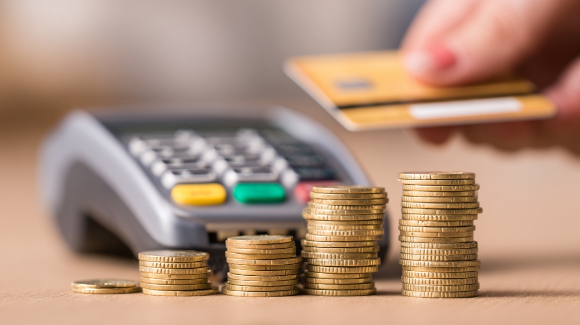 Stacks of coins beside a card payment terminal while a person holds a payment card, symbolizing digital transactions, savings, and rewards