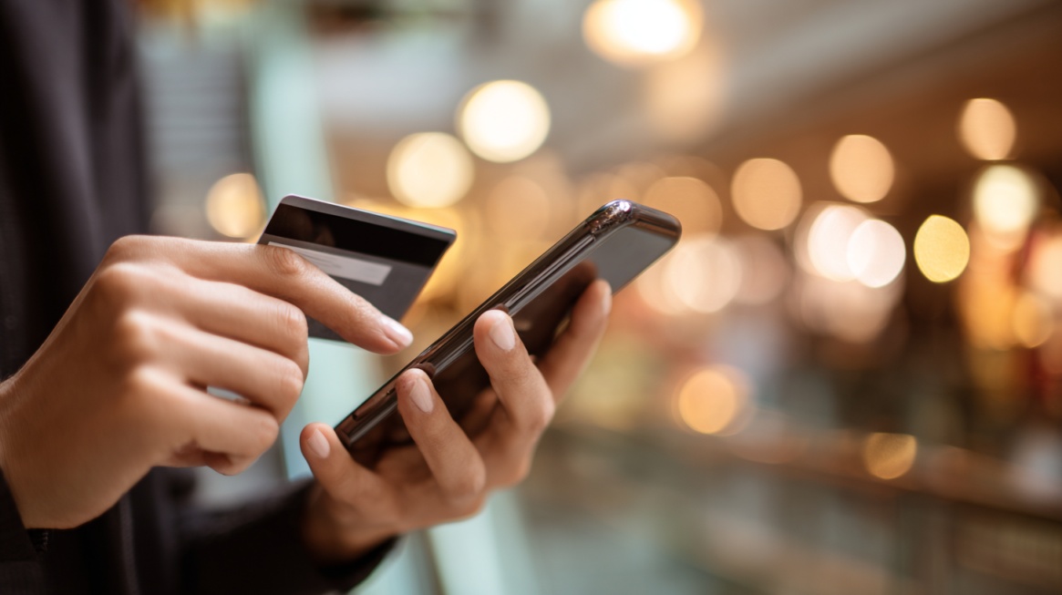 Person holding a smartphone and payment card while making a mobile purchase, with a blurred retail environment in the background
