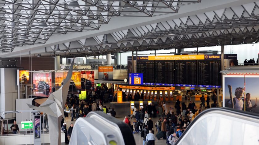Travelers walk through a busy terminal at Frankfurt Airport beneath flight information screens