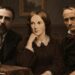 Three historical figures sit together in a dark study with books and period decor.