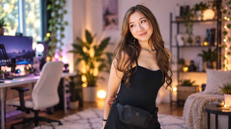 Fuslie standing in a cozy gaming room with a streaming setup in the background.