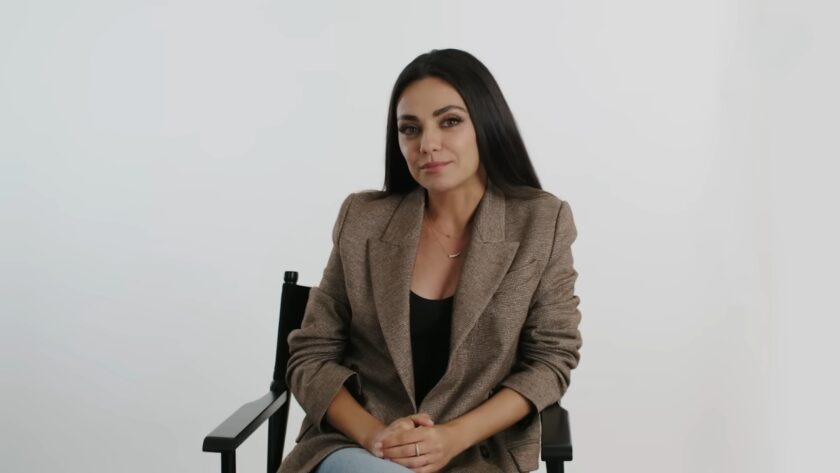 Mila Kunis in a brown blazer sits on a chair against a plain white background