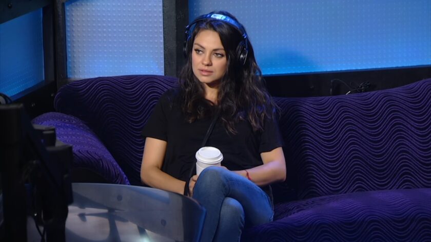 Mila Kunis with headphones sits on a purple couch and holds a coffee cup