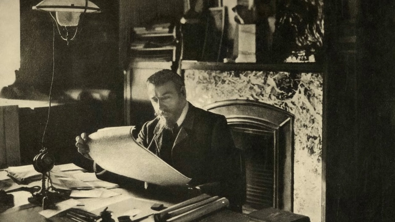 A man sits at a desk with architectural plans in a historic black-and-white photo.