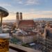Panoramic view of Munich featuring Frauenkirche, the New Town Hall, a modern car showroom, and the Bavarian Alps.