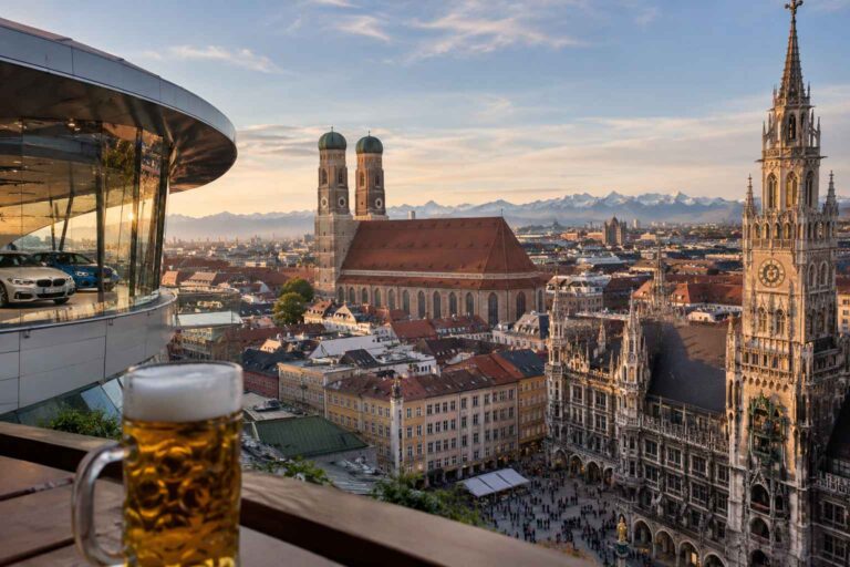 Panoramic view of Munich featuring Frauenkirche, the New Town Hall, a modern car showroom, and the Bavarian Alps.
